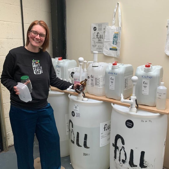 White woman wearing a black jumper with 'Food for Thought' logo (words in a jar) stands in front of big 200litre & 20 litre tubs of cleaning products. They have the word 'Fill' on them & some have descriptions hand written 'kitchen cleaner', 'rinse aid'.